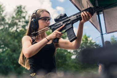 Strong powerful confident long-haired caucasian woman in a black tank top holding big black rifle with loupe preparing for a shooting competition. Medium outdoor shot. High quality photo