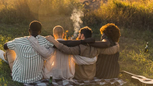 Rear view of multicultural good old friends hugging while sitting on a blanket on the grass, having a weekend picnic in nature near the river, talking and drinking beer. Sunset time. High quality
