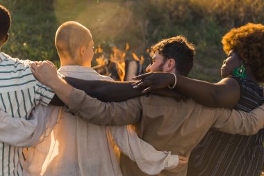 Rear view of multicultural good old friends hugging and having a weekend picnic in nature near the campfire, talking and laughing. Sunset time. High quality photo