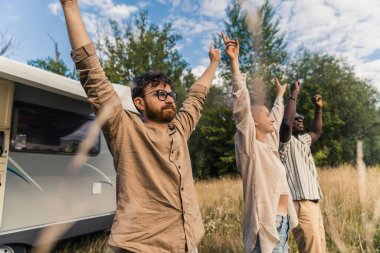 Group of happy carefree best friends, standing near their RV, raising hands and showing peace sign. Camping van road trip. High quality photo