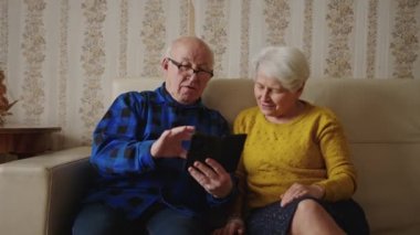 Caucasian senior man sitting with his old wife on a sofa in the living room and showing her photos on his smartphone. High quality 4k footage