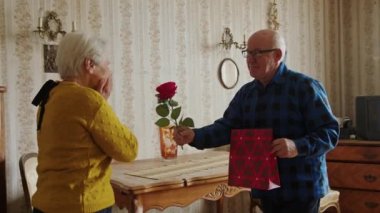 Loving caucasian elderly husband surprising his wife with a red rose and a gift bag on Valentines day. High quality 4k footage