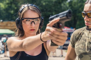 Caucasian young woman wearing protective goggles and headphones aiming handgun observed by male bearded instructor. Firearms training. Horiozntal shot. High quality photo