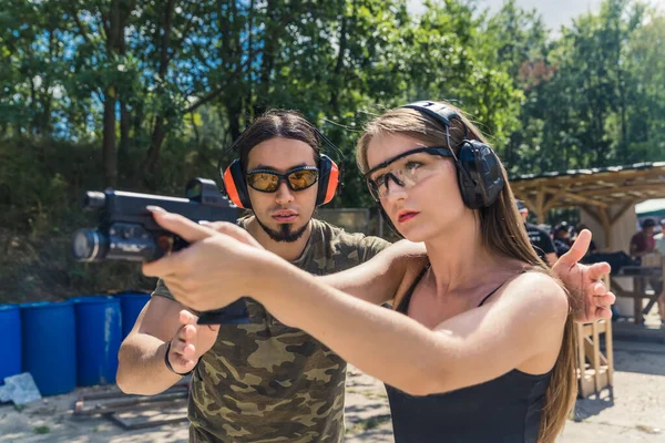 Male instructor in camo t-shirt guiding female client operating handgun. Safety headphones and goggles. Firearms training at firing range. Horizontal outdoor shot. High quality photo