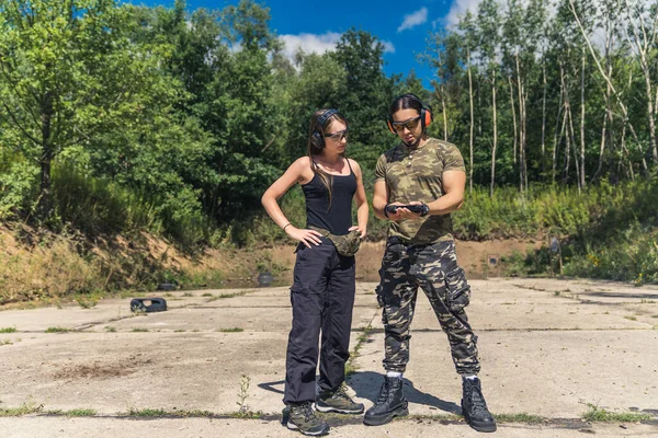 Man wearing camouflage clothing showing woman how to use handgun. Client with instructor wearing safety gear. Firearms training at outdoor shooting range. Horizontal shot. High quality photo