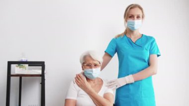Healthcare support concept. Medium indoor shot of caucasian female doctor and white-haired patient in protective masks. Healthcare professional touching elderly womans shoulders. High quality 4k
