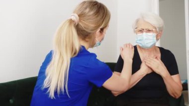 Healthcare support. Young caring caucasian blond-haired female nurse in dark blue uniform supporting her elderly lady patient. Protective masks. Pain and illness. High quality 4k footage