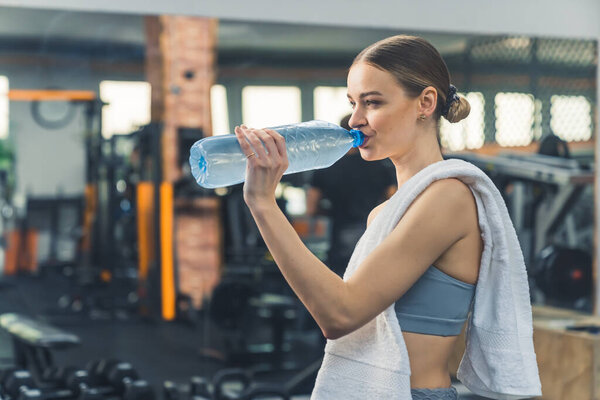 Healthy fit woman in gym wearing sportswear having towel on her shoulder, drinks water from bottle. High quality photo