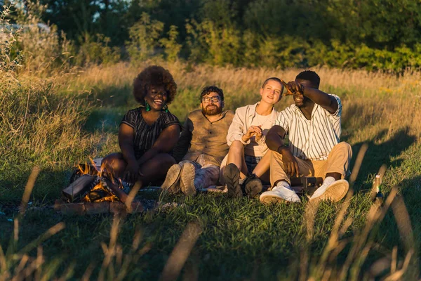 At sunset time. Happy young people having a picnic in nature, sitting together on a blanket on the grass near the campfire, laughing and looking where one of the men is pointing at. Camping at weekend