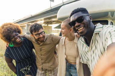 Interracial friendship. Group of happy close friends, reunited on their camping van trip, traveling through the country. Laughing and taking selfies together near the river. High quality photo