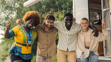 Interracial friendship on a camping van trip. Diverse people, four close friends hugging, smiling, showing a thumb up and a peace sign, taking photos together near their temporary house on wheels
