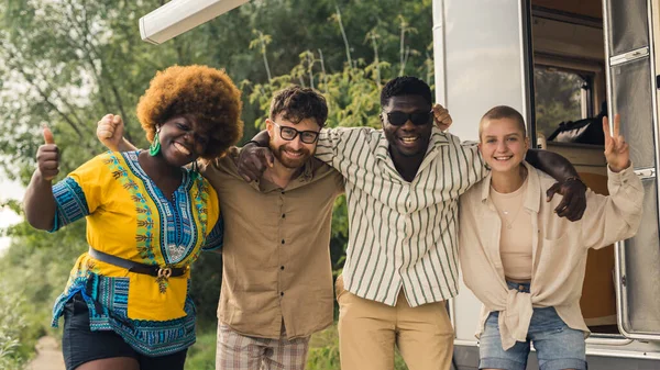 Interracial friendship on a camping van trip. Diverse people, four close friends hugging, smiling, showing a thumb up and a peace sign, taking photos together near their temporary house on wheels