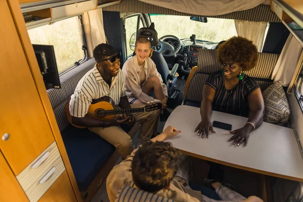 Happy interracial group of good friends on a summer road traveling, sitting inside their comfortable modern motorhome, playing the guitar, talking and laughing. RVs interior. High quality photo