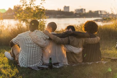 Rear view of multicultural good old friends hugging while sitting on a blanket on the grass, having a weekend picnic in nature near the river, talking, drinking beer and admiring the view. Sunset time