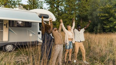 Group of happy young best friends, standing in nature, raising and holding each others hands after reaching the destination of their camping van road trip. High quality photo