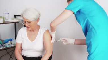 Indoor medical shot. Elderly caucasian white-haired woman in white t-shirt being vaccinated by young caucasian doctor in turquoise uniform. Virus prevention concept. High quality 4k footage