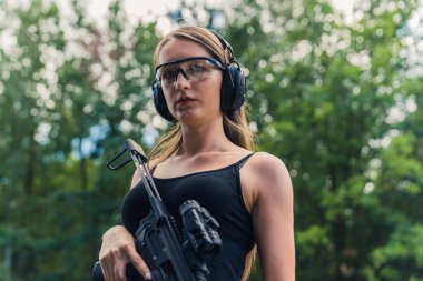 Medium closeup shot of a beautiful confident skinny caucasian girl looking at camera, wearing protective eyeglasses and headphones, and holding black rifle. Blurred background. Outdoor shot. High