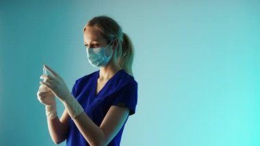 Medium closeup studio shot of a young caucasian female doctor in protective gear - face mask, white gloves, and dark blue uniform - preparing a vaccine syringe for a shot. Turquoise blue background