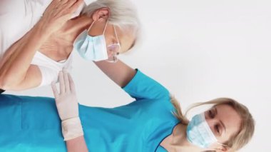 Vertical shot of two caucasian women - elderly white-haired senior in white t-shirt and blond young nurse. Healthcare worker supporting elderly people. Protective masks and gloves. High quality 4k