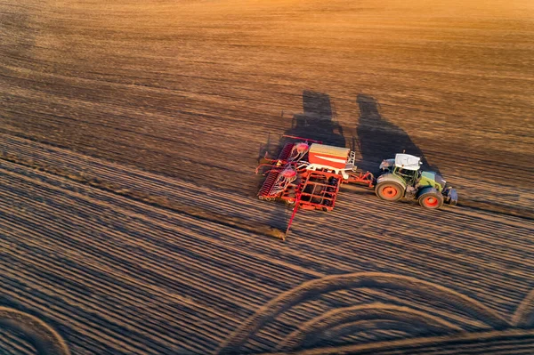 Yellow sun glare on brown field with green tractor with sowing machine driving through casting shadow. Farming and agriculture. Horizontal shot. High quality photo