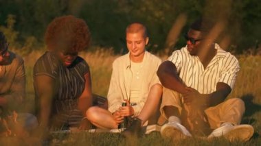 A multicultural group of friends enjoying pleasurable sunset time in nature near the campfire while sitting on the grass and talking to each other. High quality 4k footage