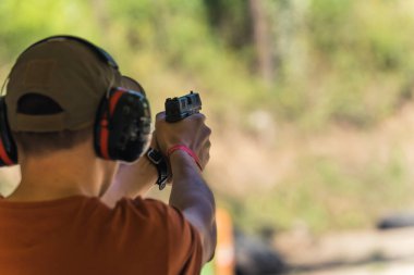 Blurred background outdoor shot. Unrecognizable man in a baseball hat, t-shirt, and protective headphones using a gun. Shooting competition concept. High quality photo