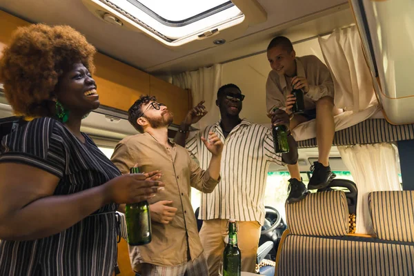 Interracial group of close friends dancing to the music and singing inside their comfortable spacious motorhome, and drinking beer. Celebrating a birthday on a camping van trip during summer. High