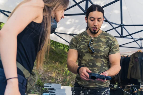Interest in weapons. Masculine muscular black-haired caucasian male gun reseller in a moro-patterned t-shirt showing his products to long-haired female customer interested in purchasing a gun. High