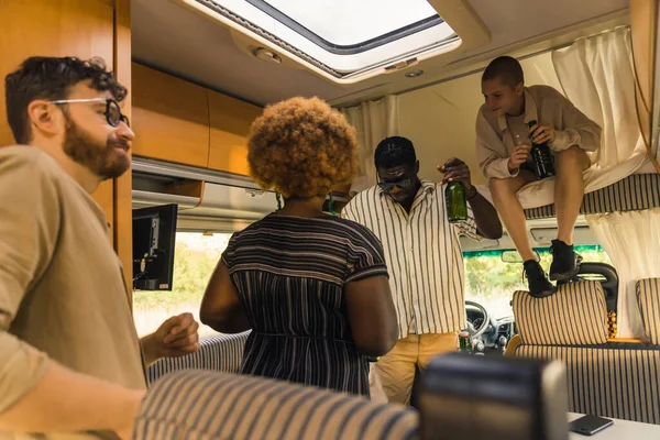 Interracial group of cheerful close friends dancing to the music and singing inside their comfortable spacious camper van, and drinking beer. Spending weekends together on a summer camping van trip