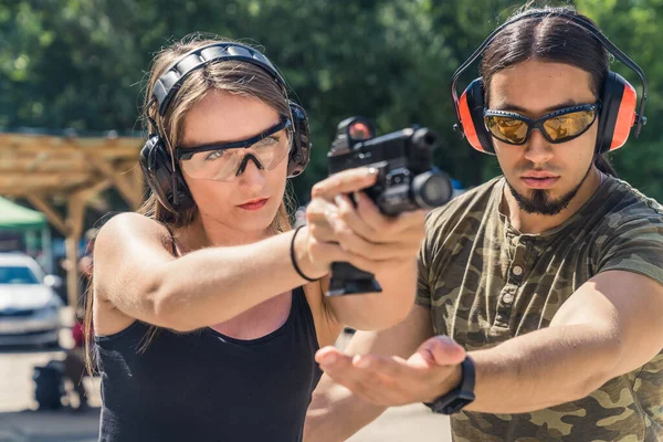 Outdoor gun range concept. Two caucasian adults - a man and a woman. Male instructor in protective gear in moro t-shirt helping out with his clients posture. High quality photo