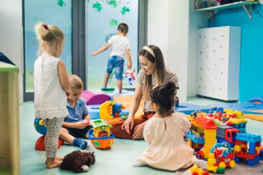 Toddlers and their nursery tutor playing with different colorful plastic toys and building blocks while sitting on the floor in a playroom. Concentration, mathematical ability, fine motor and gross