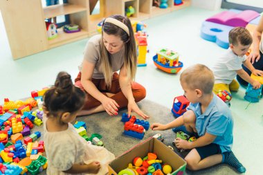 Playtime at nursery school. Toddlers with their teacher sitting on the floor and playing with building blocks, colorful cars and other toys. Fine motor and gross motor skills developing. High quality