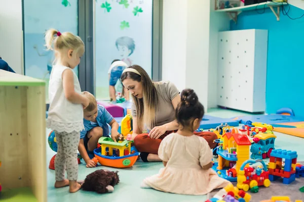 Toddlers and their nursery teacher playing with plastic building blocks, colorful cars, boats and other toys while sitting on the floor in a play room. Concentration, fine motor and gross motor skills