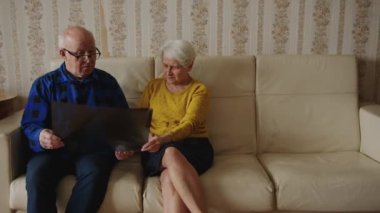 Concerned old caucasian grey-haired couple holding lungs x-ray results, looking at it and worrying about mans health while sitting on a beige leather sofa at home together. High quality 4k footage