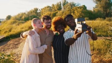 Diverse group of college friends taking funny selfies with a vintage instant camera, chilling near the river at the summer sunset time . High quality 4k footage