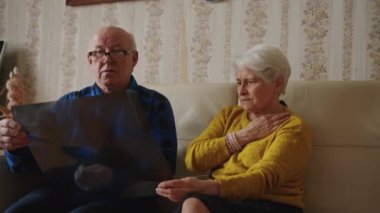 Concerned old caucasian grey-haired married couple holding x-ray results, scan of a mans lungs, and worrying about health while sitting on a beige leather sofa at home together. High quality 4k