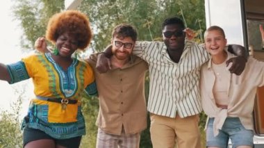 Interracial friendship on a camping van trip. Diverse people, four close friends hugging, smiling, showing a thumb up and a peace sign, taking photos together near their temporary house on wheels