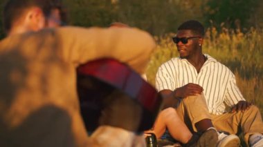 Group of happy young people having a weekend picnic in nature, sitting together on the grass near the campfire, talking and laughing. Sunset time. High quality 4k footage