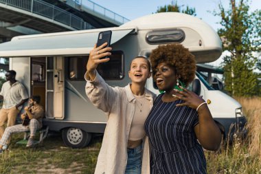 Interracial friendship. Two young diverse girls taking a selfie on a phone together near their camper van. The boys sitting on the stairs and playing the guitar in the background. Reunited on their