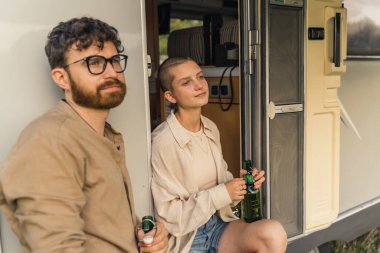 Young caucasian girl and a man holding bottles of beer and resting against their camping van doors, thoughtfully looking away. Motorhome road trip. High quality photo