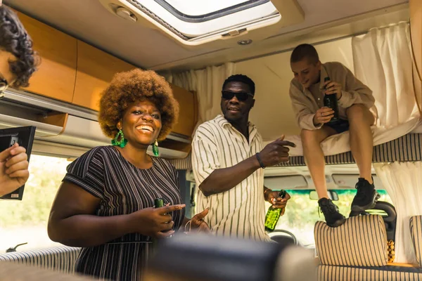 Interracial group of close friends dancing to the music inside their comfortable spacious motorhome and drinking beer. One of the girls sitting on an over cab bed. Celebrating their reunion by a