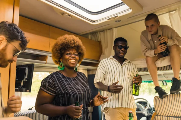 Interracial group of close friends traveling in a camping van during summer, dancing to the music inside their comfortable spacious motorhome and drinking beer. One of the girls sitting on an over cab