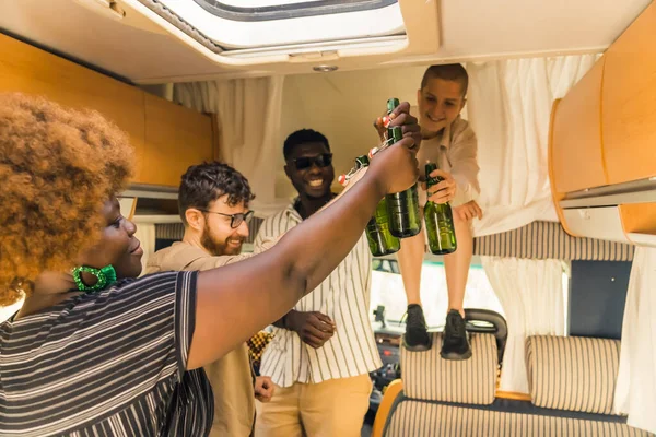 Interracial group of cheerful close friends toasting with beer bottles, having drinks to celebrate a special occasion inside their comfortable spacious camper van. Spending weekends together on a