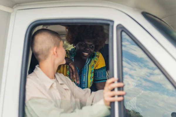 Young caucasian pretty woman with a shaved head sitting near the open window of a camping van and discussing something with her African female friend on the back seat. High quality photo