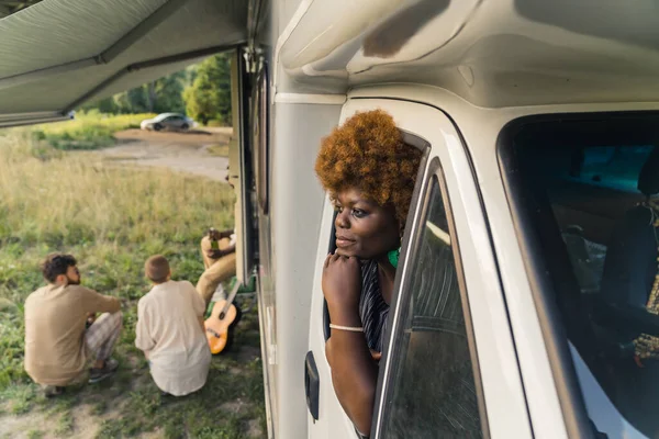 Thoughtful dark-skinned African young woman with kinky hair leaning out of the window of a camper van and her friends sitting on the grass next to her at the end of their road trip. High quality photo