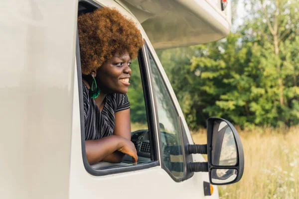 Dark-skinned smiling African young woman with kinky hair leaning out of the window of a camper van and admiring peaceful nature around at the end of her road trip. High quality photo