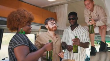 Interracial group of cheerful close friends toasting with beer bottles, having drinks to celebrate a special occasion inside their comfortable spacious camper van. Spending weekends together on a