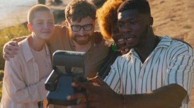 Multi-ethnic group of best friends creating memories by taking funny photos with a vintage polaroid, chilling near the river at the summer sunset time . High quality 4k footage