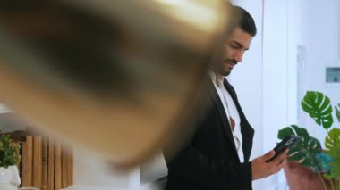 Hispanic smiling businessman seen from behind a gold lamp in white shirt and blazer making a call on smartphone working from home. Indoor. High quality 4k footage