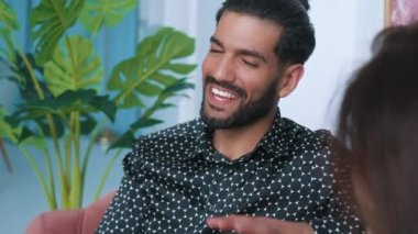 ark-haired man with beard enthusiastically talking to his girlfiend smiling sitting on the couch together in the living room. Young couple. Indoor. High quality 4k footage